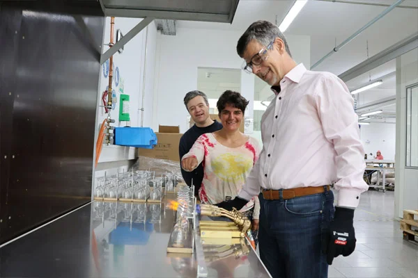 Ein Mann mit Schutzbrille arbeitet und zwei Personen stehen im Hintergrund.