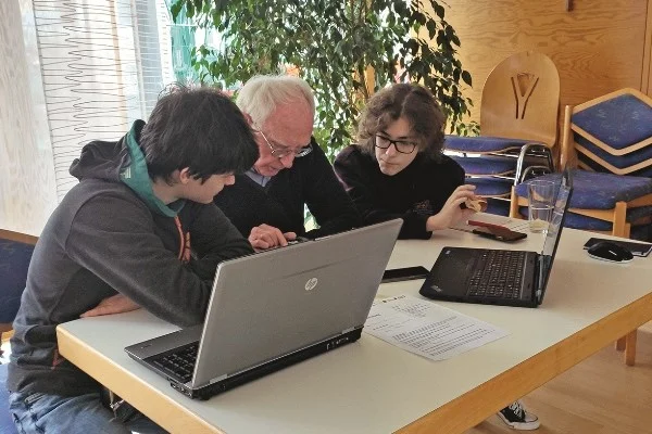  Das Bild zeigt zwei Jungen die gerade einem älteren Herrn helfen den PC zu verstehen
