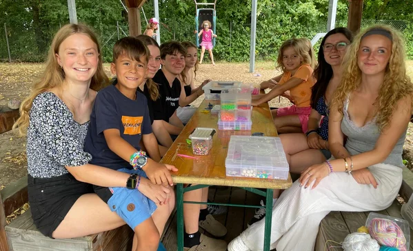 Ferienbetreuung Olympia Kinder und junge Erwachsene sitzen auf einer Bank in der Natur und lächeln in die Kamera.