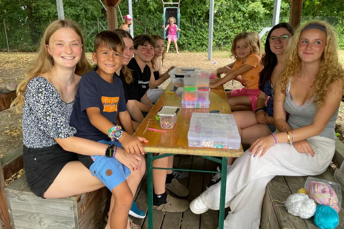 Ferienbetreuung Olympia Kinder und junge Erwachsene sitzen auf einer Bank in der Natur und lächeln in die Kamera.