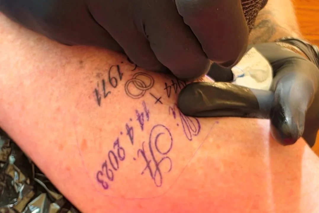 Tattoo Nahaufnahme eines Tätowierers mit schwarzen Handschuhen, der mit einer Tätowiermaschine Schrift und Zahlen auf dem Unterarm tätowiert.