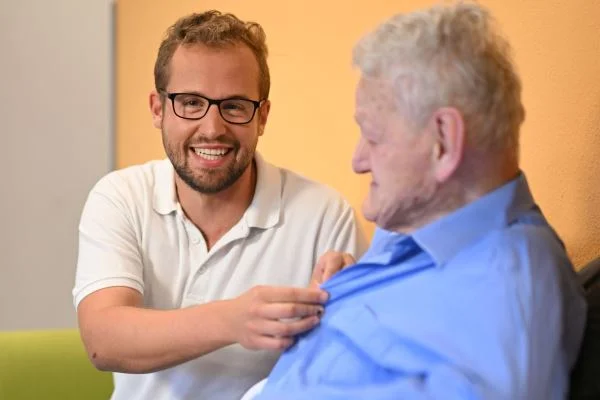 Auf dem Bild sieht man einen Mann in weißem Polo der einem älteren Mann das Hemd zuknöpft