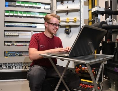 Das Bild zeigt einen jungen Mann an einem PC vor einer Maschine