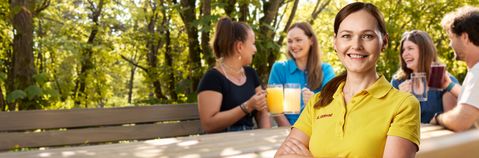 Im Vordergrund ist eine Mitarbeiterin in Arbeitskleidung zu sehen. Im Hintergrund sieht man vier junge Personen, die an einem Tisch im Garten sitzen.