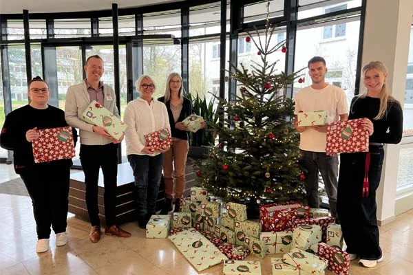Wunschbaum Viele Personen mit Geschenken stehen um einen Christbaum.