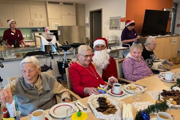 Auf dem Bild sieht man mehrere ältere Menschen die gemeinsam an einem Tisch sitzen und Kuchen essen. Um sie herum stehen Mitarbeitende die als Nikolaus verkleidet sind