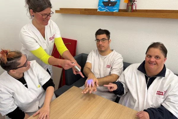 Das Bild zeigt eine Gruppe am Tisch und eine Frau mit einem UV-Gerät