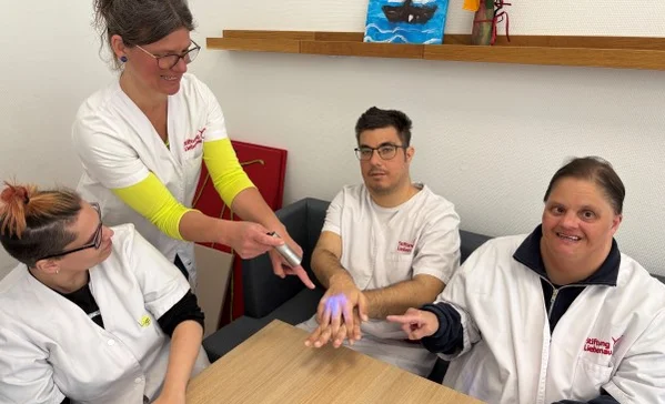 Das Bild zeigt eine Gruppe am Tisch und eine Frau mit einem UV-Gerät