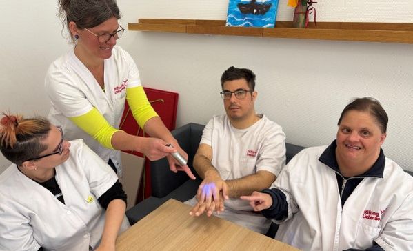 Das Bild zeigt eine Gruppe am Tisch und eine Frau mit einem UV-Gerät