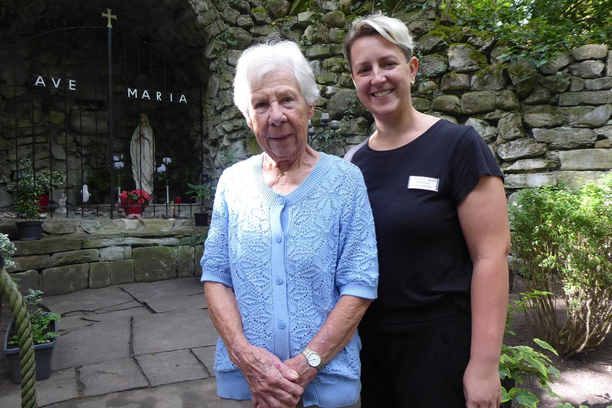 Zwei lächelnde Frauen, eine ältere in heller blauer Strickjacke und eine jüngere in schwarzem Oberteil mit Namensschild, stehen nebeneinander vor einer steinernen Mariengrotte mit einer Madonnenstatue und der Aufschrift AVE MARIA