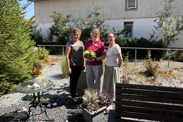 Drei lächelnde Frauen stehen nebeneinander in einem sonnigen Garten; die mittlere hält einen Blumenstrauß.