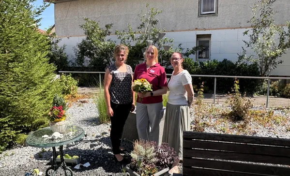 Drei lächelnde Frauen stehen nebeneinander in einem sonnigen Garten; die mittlere hält einen Blumenstrauß.