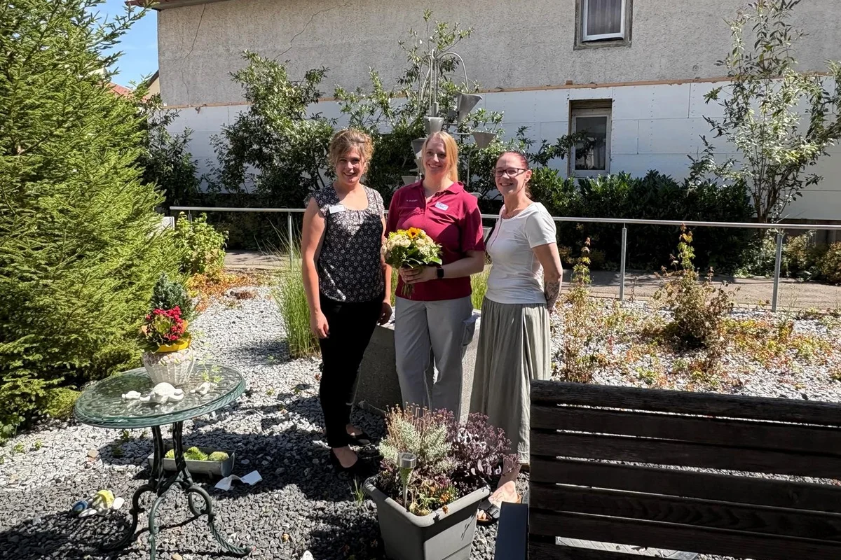St. Wunibald Scheer Miriam Stump Drei lächelnde Frauen stehen nebeneinander in einem sonnigen Garten; die mittlere hält einen Blumenstrauß.