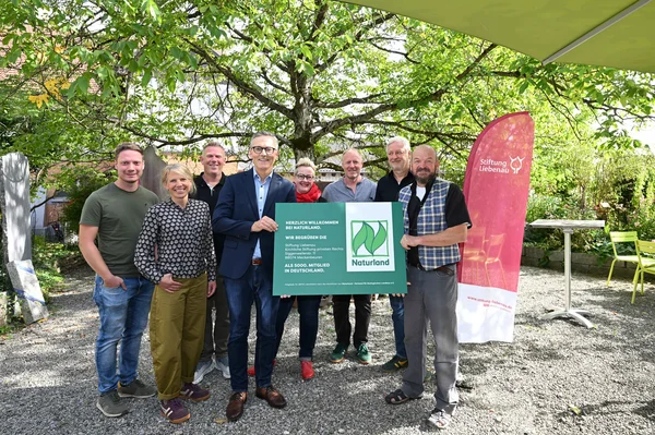 Neun lächelnde Erwachsene stehen in einem Garten unter Bäumen und halten gemeinsam ein grünes Schild mit Naturland-Logo; seitlich steht ein roter Werbebanner der Stiftung Liebenau.