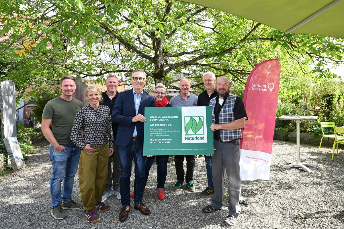 5.000ster Naturland Betrieb in Deutschland Neun lächelnde Erwachsene stehen in einem Garten unter Bäumen und halten gemeinsam ein grünes Schild mit Naturland-Logo; seitlich steht ein roter Werbebanner der Stiftung Liebenau.