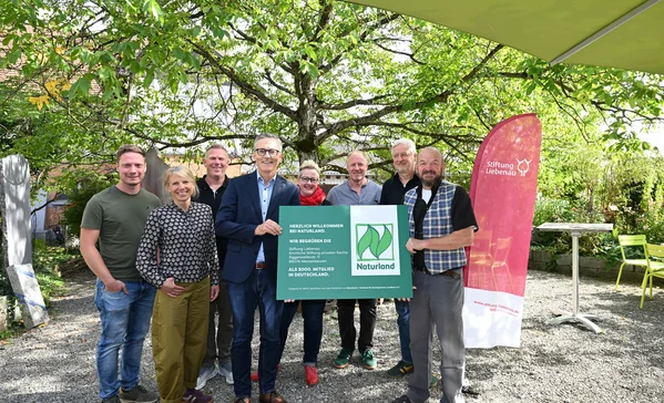Neun lächelnde Erwachsene stehen in einem Garten unter Bäumen und halten gemeinsam ein grünes Schild mit Naturland-Logo; seitlich steht ein roter Werbebanner der Stiftung Liebenau.
