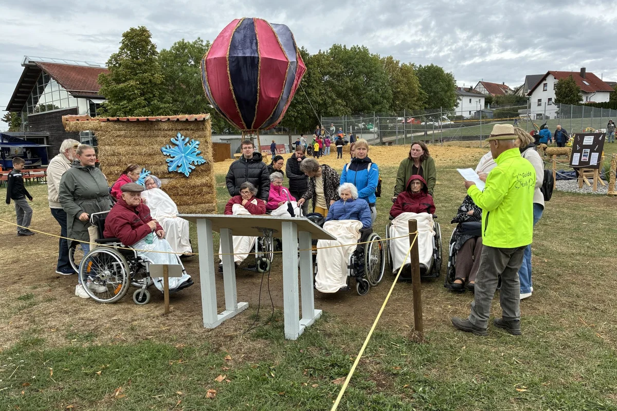 Bewohnerausflug Strohpark Gruppe mehrerer Menschen in Rollstühlen mit Betreuern bei einer Freiluftveranstaltung auf einer Wiese; Dekoration aus Heuballen und großer bunter Heißluftballon-Attrappe, eine Person in gelber Jacke liest vor.