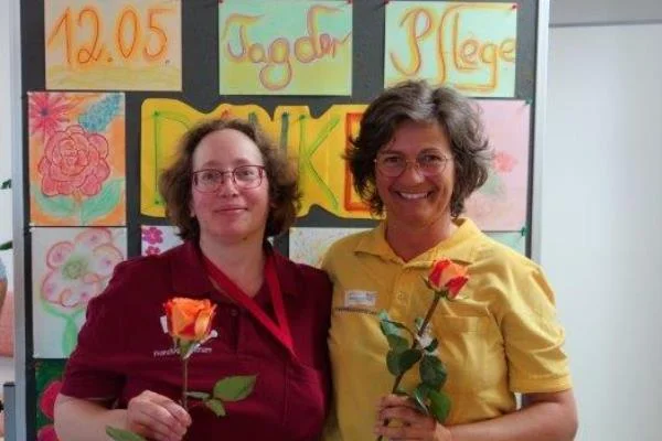 Aktionen zum Tag der Pflege Das Bild zeigt zwei Frauen vor einer Bildercollage mit je einer Rose in der Hand