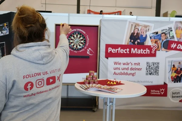 Eine Frau zielt mit einem Dartpfeil auf eine Dartscheibe an einem Messestand.