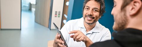 Das Bild zeigt zwei Männer mit einem Tablet auf einem Gang und verlinkt zu Jobs in der Gesundheit