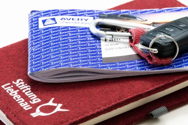 Das Bild zeigt ein Stiftung Liebenau Block, einen Autoschlüssel und ein weiteres blaues Heft.