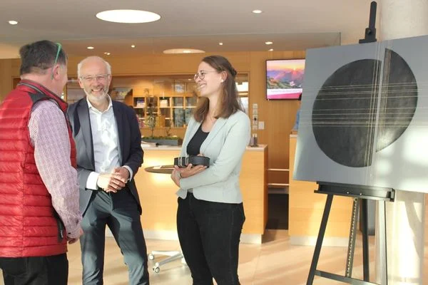 Zwei Männer und eine Frau Stehen vor einem Ausstellungsstück, das eine Art Kreis zeigt und reden lächelnd miteinander.