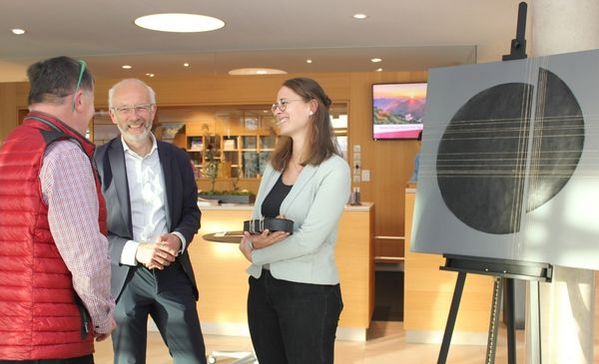 BM Smigoc Zwei Männer und eine Frau Stehen vor einem Ausstellungsstück, das eine Art Kreis zeigt und reden lächelnd miteinander.