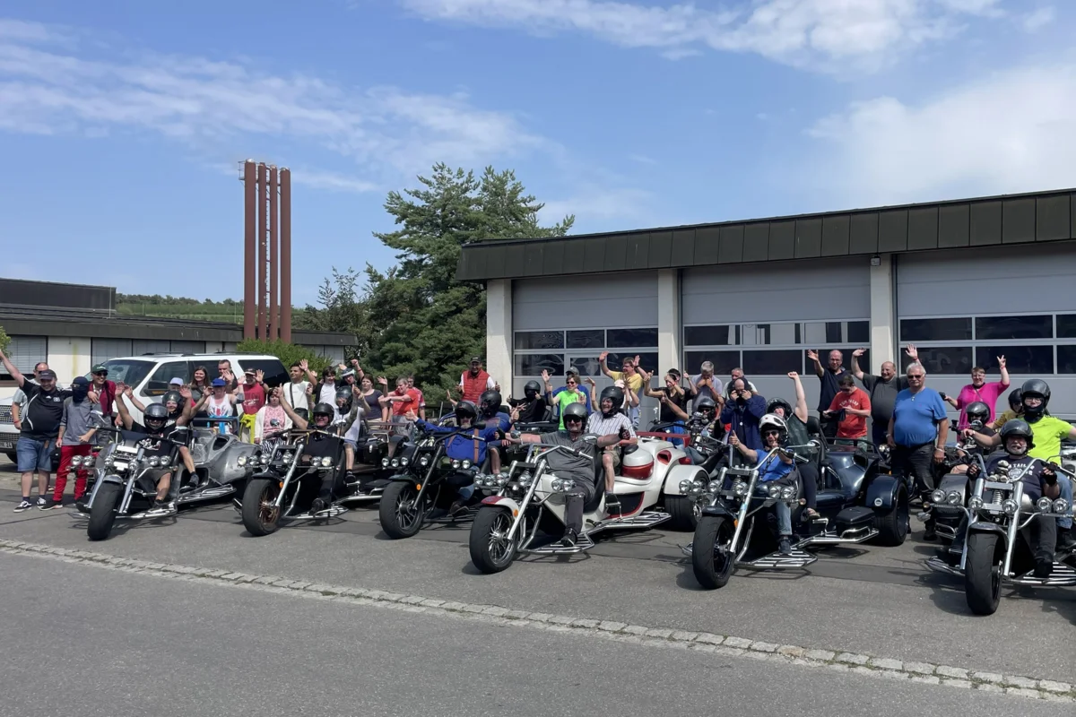 Stiftung Liebenau Trike Große Gruppe von Motorradfahrern und Beifahrern mit zahlreichen Motorrädern und Trikes vor einem Garagengebäude, lächelnd und mit erhobenen Händen winkend.