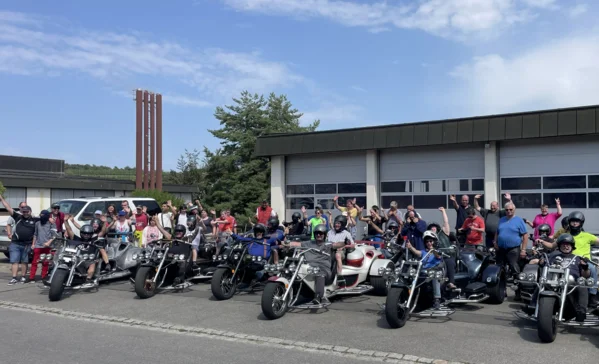 Große Gruppe von Motorradfahrern und Beifahrern mit zahlreichen Motorrädern und Trikes vor einem Garagengebäude, lächelnd und mit erhobenen Händen winkend.