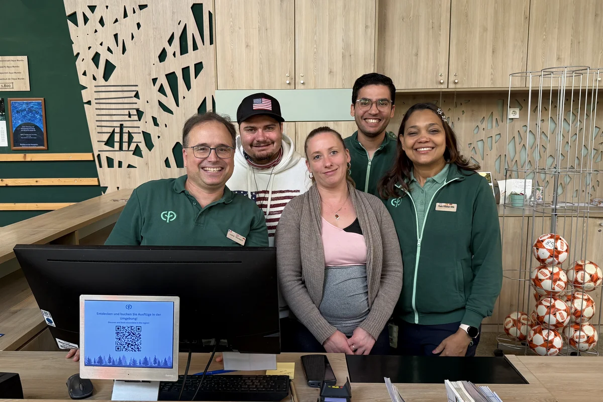 Team Rezeption Fünf lächelnde Mitarbeitende stehen eng nebeneinander hinter einer Empfangstheke; drei tragen grüne Dienstkleidung, vor ihnen ein Computermonitor.