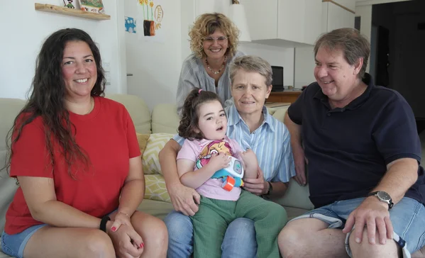 Multigenerationenfamilie auf Sofa: Eine ältere Frau hält ein kleines Mädchen auf dem Schoß, zwei Erwachsene sitzen zu beiden Seiten, eine Frau steht lächelnd dahinter.