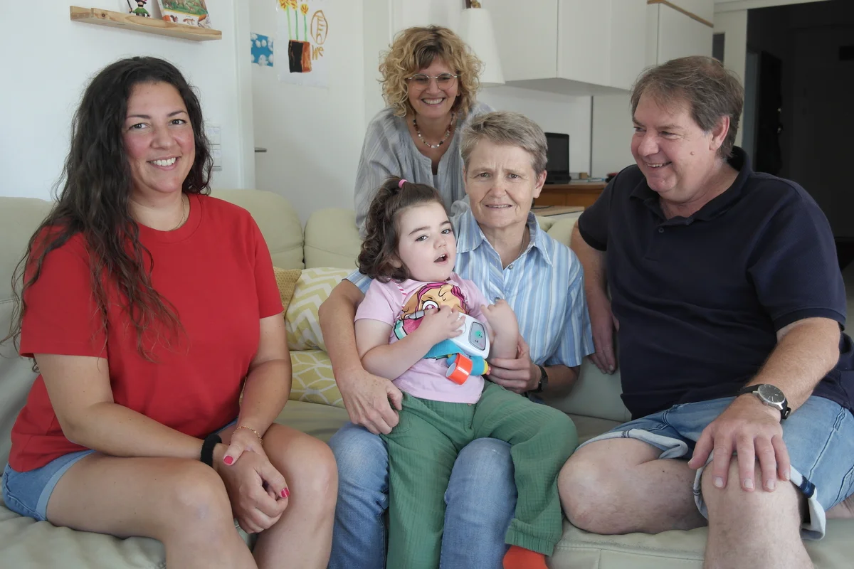 Familienunterstützender Dienst Multigenerationenfamilie auf Sofa: Eine ältere Frau hält ein kleines Mädchen auf dem Schoß, zwei Erwachsene sitzen zu beiden Seiten, eine Frau steht lächelnd dahinter.