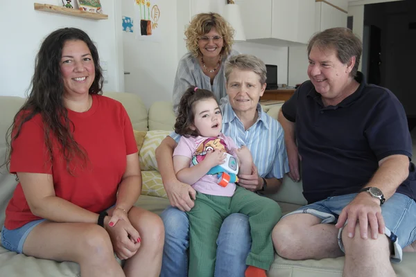 Multigenerationenfamilie auf Sofa: Eine ältere Frau hält ein kleines Mädchen auf dem Schoß, zwei Erwachsene sitzen zu beiden Seiten, eine Frau steht lächelnd dahinter.
