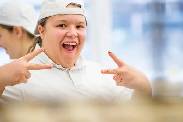Das Bild zeigt eine Person die weiß gekleidet ist und eine weiße Mütze auf dem Kopf trägt und vier Finger in die Kamer zeigt 
