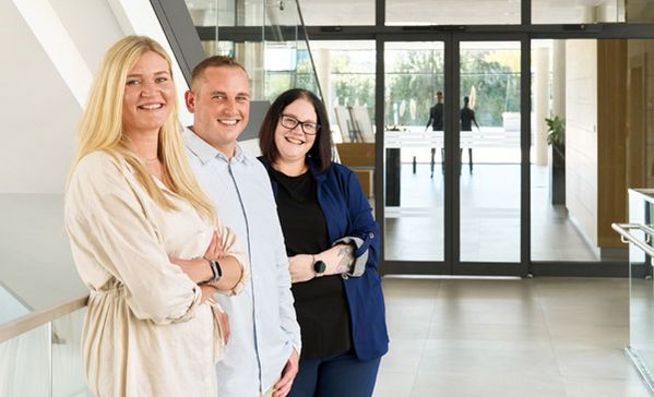 Zwei Frauen und ein Mann stehen in einem Flur und lächeln in die Kamera.