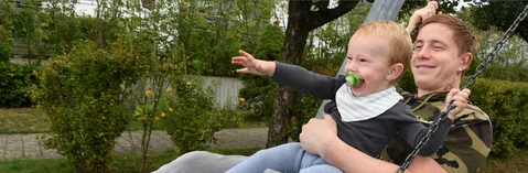 Das Bild zeigt einen jungen Mann mit einem Baby auf einer Schaukel