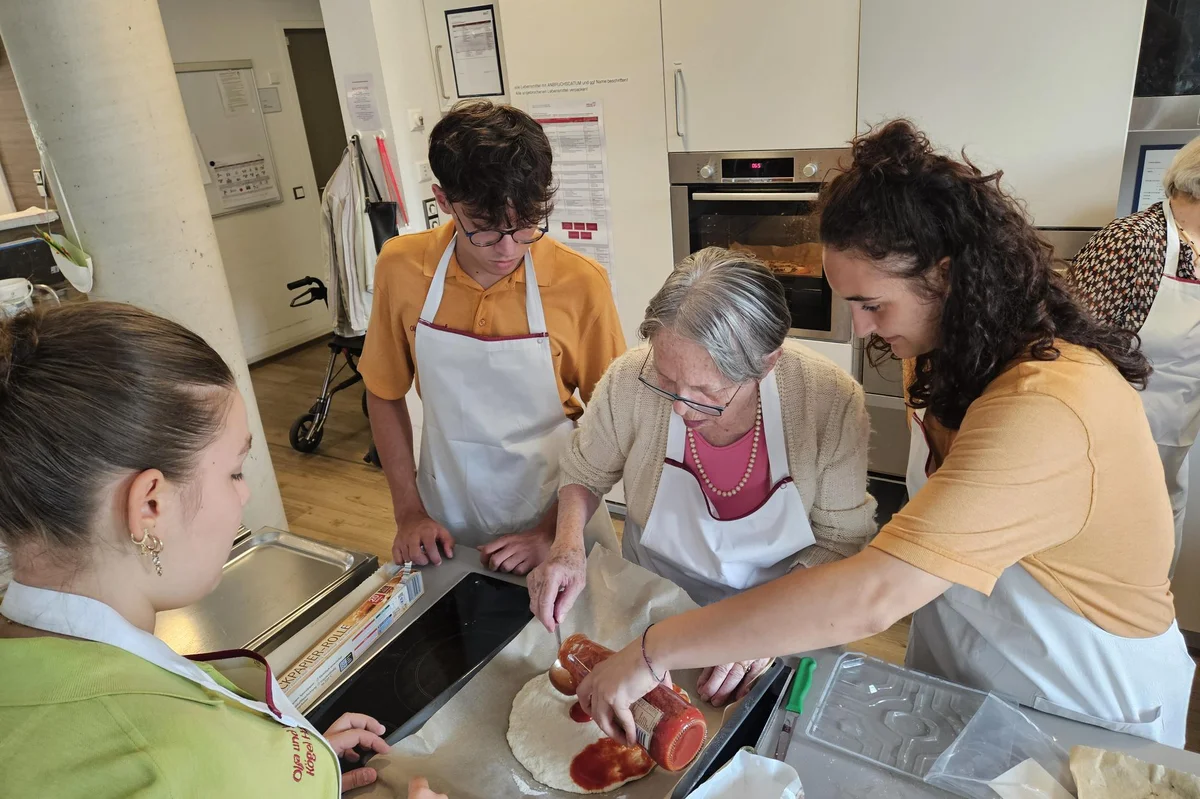 Vier Personen stehen in der Küche und bestreichen eine Pizza mit Tomatensauce.