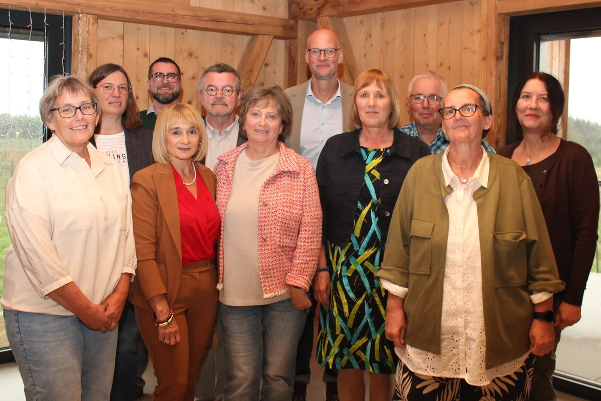 Jubilarehrung Zwölf Erwachsene posieren in einem hellen, holzgetäfelten Raum, stehend in zwei Reihen, lächelnd und gut gekleidet.