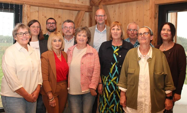 Zwölf Erwachsene posieren in einem hellen, holzgetäfelten Raum, stehend in zwei Reihen, lächelnd und gut gekleidet.
