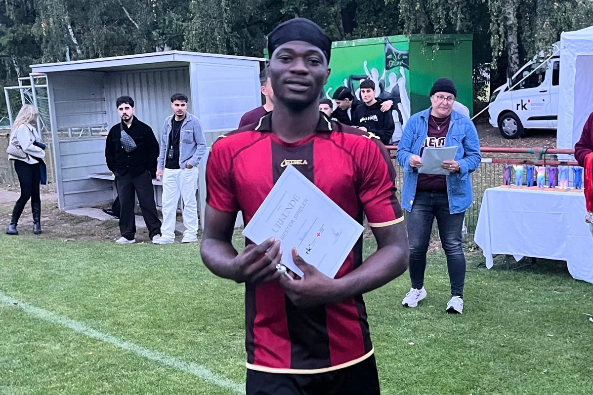 Bouba - Spieler des Turniers Person in rot-schwarzem Fußballtrikot läuft über Rasen und hält eine Urkunde, im Hintergrund Zuschauer und ein Tisch mit Pokalen.