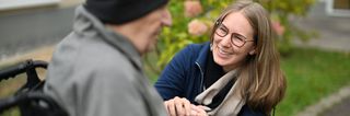 Das Bild zeigt eine junge Frau, die einem älteren Mann im Rollstuhl die Hand hält