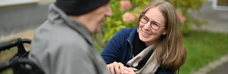 Das Bild zeigt eine junge Frau, die einem älteren Mann im Rollstuhl die Hand hält