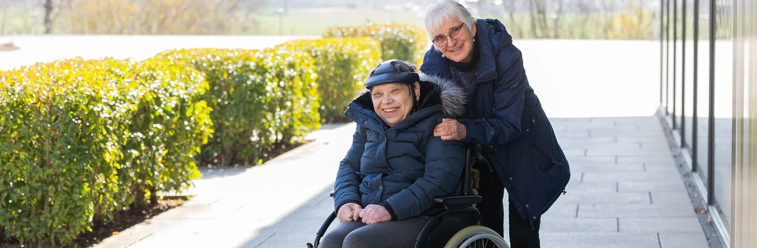 Das Bild zeigt eine Frau die eine andere Frau im Rollstuhl schiebt und beide Frauen lächeln in die Kamera 