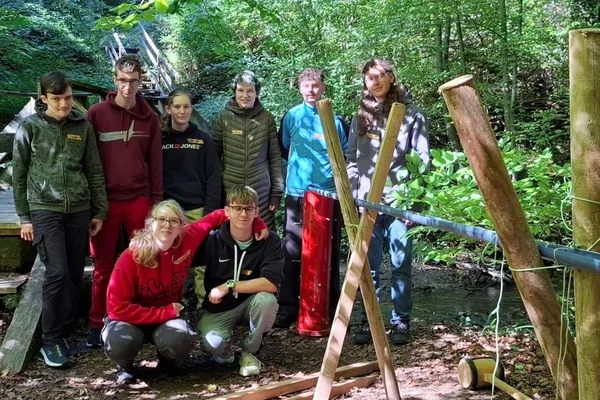 Acht Jugendliche posieren in einem Wald neben einer selbstgebauten Holzkonstruktion mit Seilen, offenbar bei einer Outdoor-Teamaktivität.
