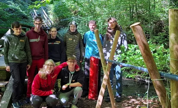 Acht Jugendliche posieren in einem Wald neben einer selbstgebauten Holzkonstruktion mit Seilen, offenbar bei einer Outdoor-Teamaktivität.