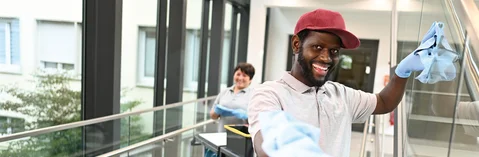 Das Bild zeigt einen Mann der in die Kamera schaut und dabei in einem Gebäude ein Treppengeländer putzt, außerdem verlinkt das Bild zum Arbeitgeber-Video des Service