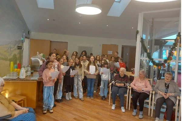 Das Bild zeigt eine Gruppe von Kindern die Lieder singen. Sie halten Liedzettel in der Hand um am Rand sitzen mehrere ältere Menschen und hören zu.