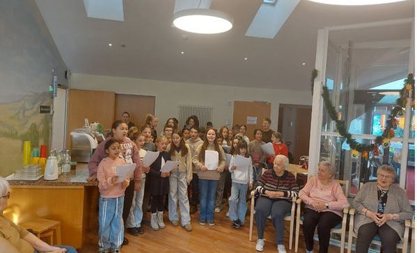 Das Bild zeigt eine Gruppe von Kindern die Lieder singen. Sie halten Liedzettel in der Hand um am Rand sitzen mehrere ältere Menschen und hören zu.