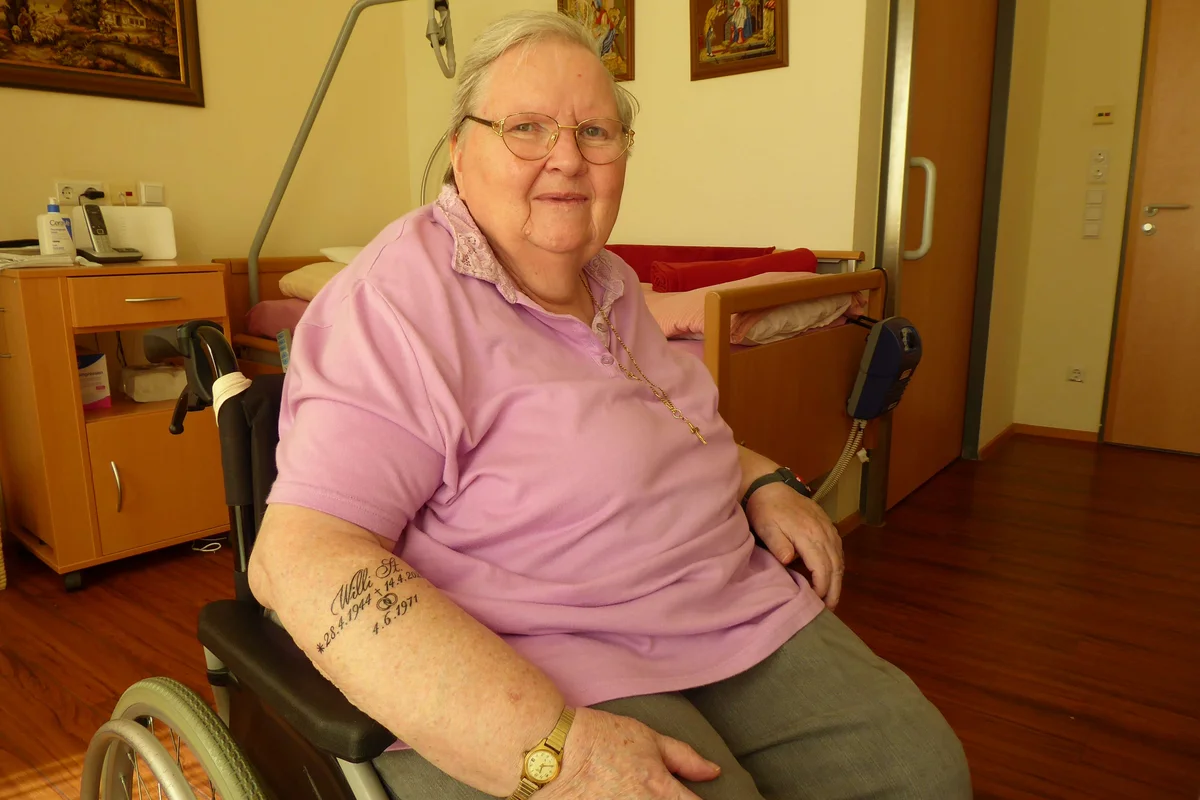 Tattoo Ältere Frau sitzt im Rollstuhl in einem Zimmer, trägt rosa Poloshirt und Brille, lächelt in die Kamera; Uhr am Handgelenk und Tattoo am Unterarm sichtbar.