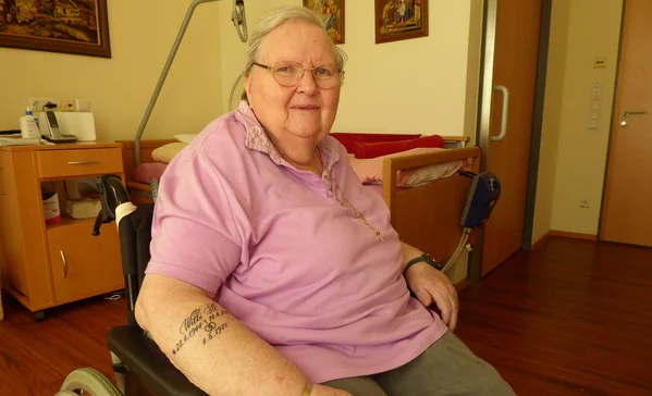 Ältere Frau sitzt im Rollstuhl in einem Zimmer, trägt rosa Poloshirt und Brille, lächelt in die Kamera; Uhr am Handgelenk und Tattoo am Unterarm sichtbar.